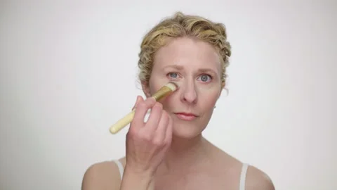 Makeup Demonstration: Curly blonde blue eyed model brushes foundation under eyes Video stock 193831006