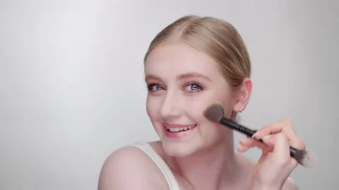 Makeup Tutorial: Beautiful blonde model happily applies blush looking in camera 스톡 동영상 193709951