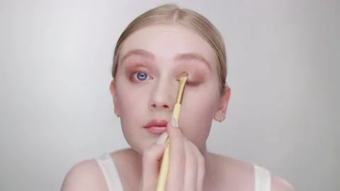 Makeup Tutorial: Pretty blonde model applies eye shadow to eye crease in studio Stock Footage 193703407