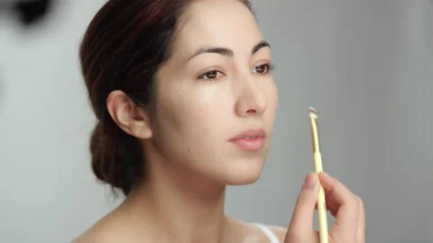 Makeup tutorial: Side view, lovely brunette woman applies foundation with brush 스톡 동영상 193853717