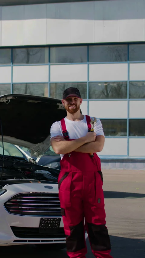 Male auto technician in red overalls stands confidently beside a white car with Stock Footage 320958853
