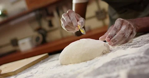 Male baker noticing proofed dough, scoring loaf with yellow tool on floured wood Stock Footage 341394448