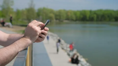The male on a bench, and scrolls through the pictures in the phone, on promenade Stock Footage 253806730