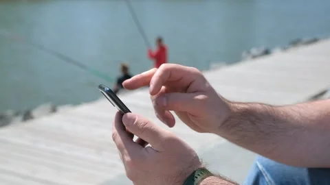 The male on a bench, and scrolls through the pictures in the phone, on promenade Stock Footage 257460809