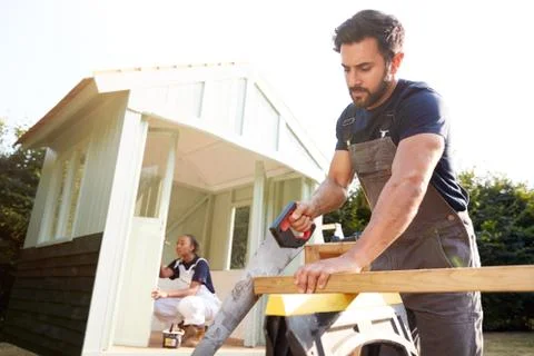 Male Carpenter With Female Apprentice Sawing Wood To Build Outdoor Summerhouse Stock Photos