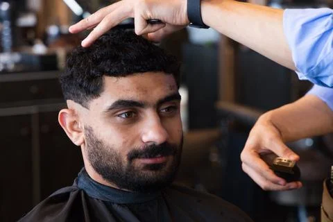 Male client getting haircut while male barber in blue shirt trimming with Stock Photos