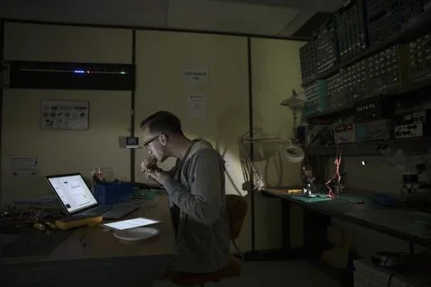 Male computer programmer working at laptop eating in dark workshop Foto stock