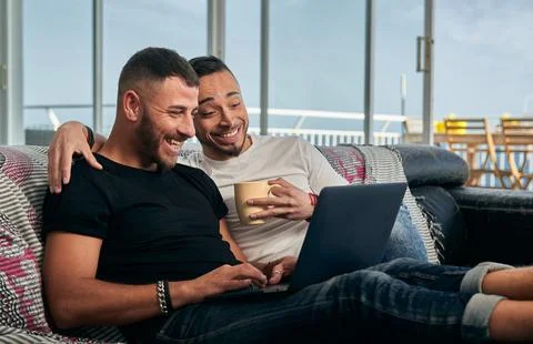 Male couple smiling while using a laptop sitting on the couch at home. Stock Photos
