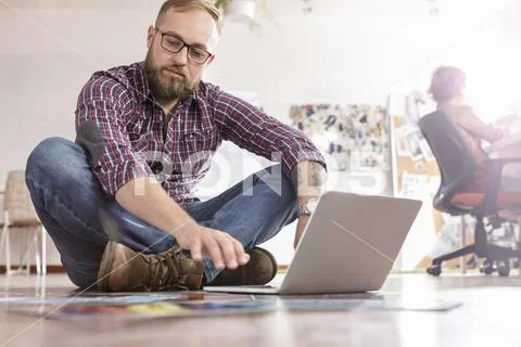 Male design professional reviewing proofs at laptop on office floor Stock Photos