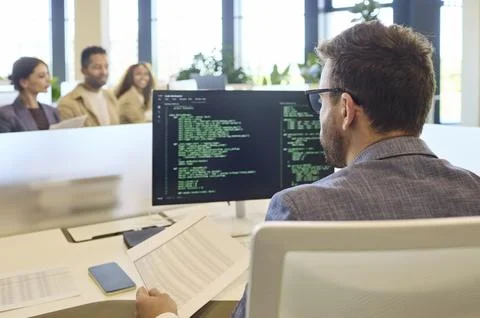 Male developer coding software at computer monitor in modern office workspace Stock Photos