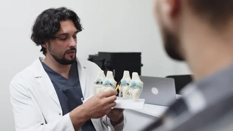 Male doctor explaining knee joint anatomy to patient using anatomical models in Stock Footage 339980416
