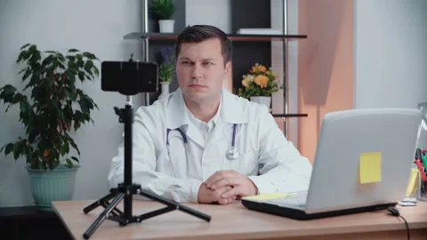 A male doctor, sitting at a table, leads his video blog, records himself Stock Footage 152024937