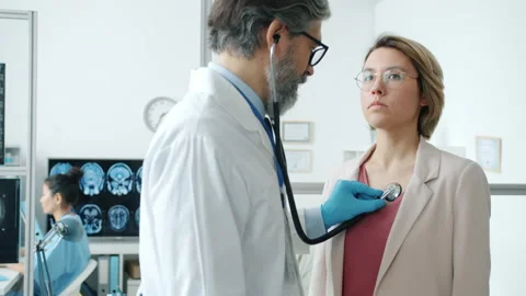 Male doctor using stethoscope while female patient breathing during medical exam Stock Footage