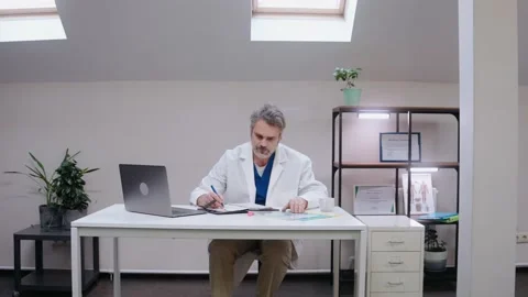 Male elderly doctor writes notes sitting at the desk, wearing medical gown Vídeo Stock 166665418