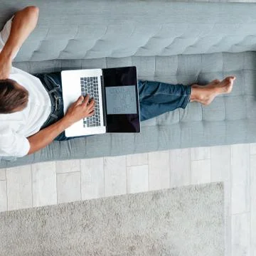 Male freelancer is working on a laptop while lying on the couch . Stock Photos