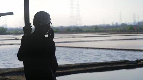 Male Indian worker talking on a mobile phone silhouetted against salt pan flats 스톡 동영상 337092864