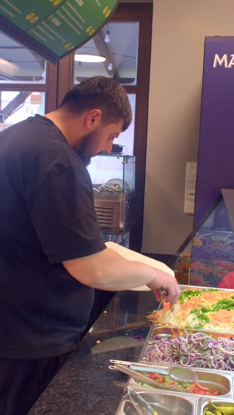 Male kitchen worker assembling flatbread behind glass service counter Vidéo 336535002