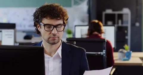 Male office worker sitting at the computer and working with printed documents in Stock Footage 82445599