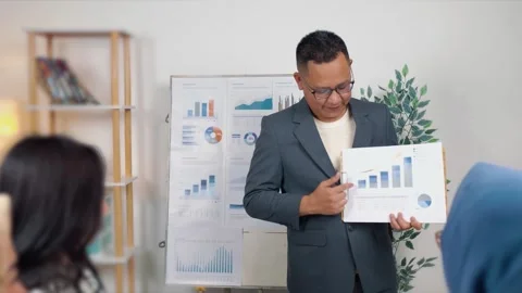 Male office worker standing in front of a team, holding and pointing at a g.. Stock Footage 344090999