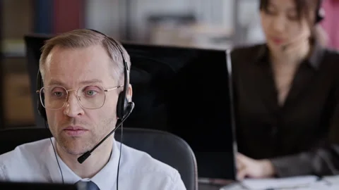 Male Operator Speaking via Headset Working in Call Center with Colleagues 스톡 동영상 335749605