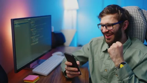 A male programmer looks at the screen of a mobile phone sitting at his desk and Stock-Footage 228940570