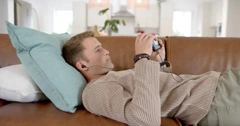 Male reclining on sofa, propping head on teal pillow, adjusting device to check Stock Footage 344405423