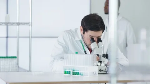 Male researcher looking through microscope and hold test tube of green solution Stock Footage 107591890