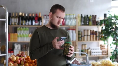 Male shopper scans a QR code on a label of canned cucumbers in a supermarket Stock Footage 329543216