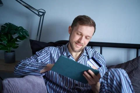 Male sitting on bed using tablet for shopping, purchasing with credit card Stock Photos