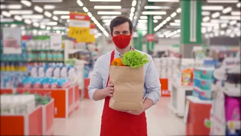 Male store courier in face mask standing in supermarket with vegetables in paper Stock Footage 147995062