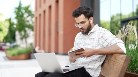 Male student studies online using laptop taking notes while sitting on a bench  Stock Footage 306300794