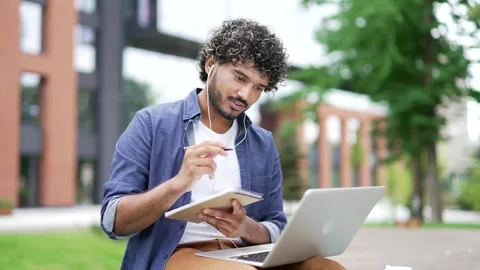 Male student studies online using laptop taking notes while sitting on a bench  Stock Footage 317710488