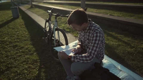 Male student taking notes in a notebook sitting in the park near his bicycle Stock Footage 222401235