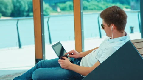 Male in sunglasses using tablet pc sitting on outdoor bench against the river Stock Footage 64779871