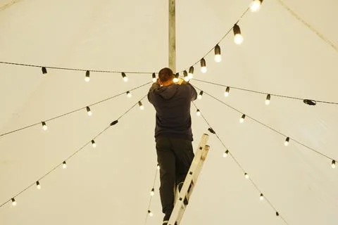 Male technician adjusting decorative string lights on a ladder inside a large Stock Photos