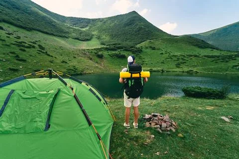 A male tourist with a backpack and a rubber mat stands near a tent by the lak Stock Photos