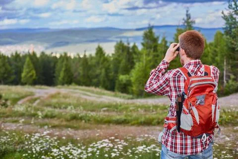 Male traveler watching binoculars into the distance against a forest and a cl Stock Photos