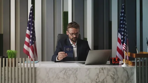 Male U.S. embassy worker processing American passport data on laptop in office Stock Footage 327784613