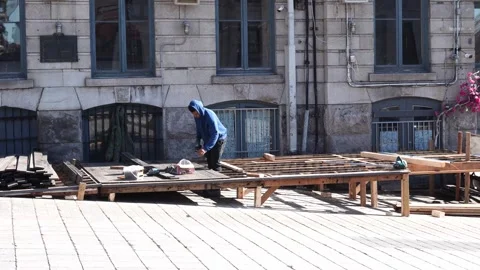 A male worker in a blue hoodie constructs a wooden platform or outdoor terrace Stock Footage 334330118