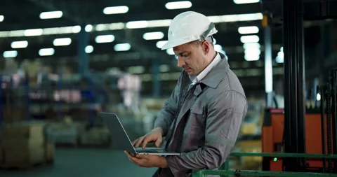 Male worker in gray uniform using laptop beside forklift in warehouse, focusing Stock Footage 307854206