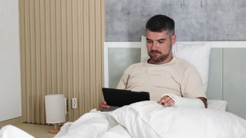 Man adapts to life with broken arm while using tablet in cozy bedroom. Stock Footage 289551908