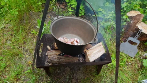 Man adding chopped bacon into cauldron over campfire for outdoor picnic coo.. 動画素材 343550803