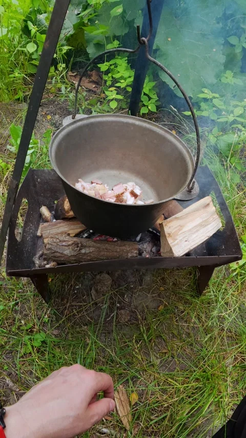 Man adding chopped bacon into cauldron over campfire for outdoor picnic coo.. Stock Footage 348091087