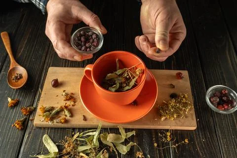 A man is adding dried fruit to an orange mug filled with various herbs. Mor.. Stock Photos