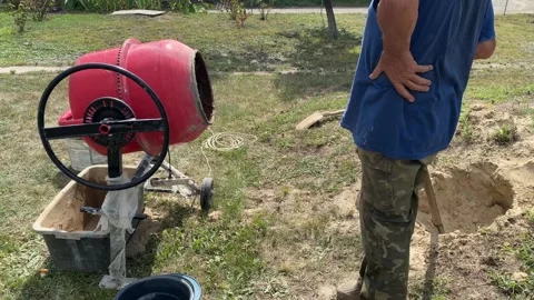 Man adding water to red concrete mixer with shovel and clay bucket nearby Stock Footage 316539391