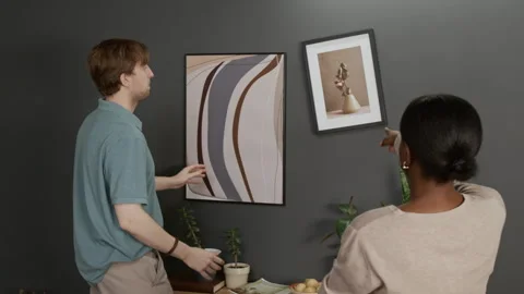 Man Adjusting Alignment of Picture Frames on Wall, Girl Watching and Guiding Him Stock Footage 335157124