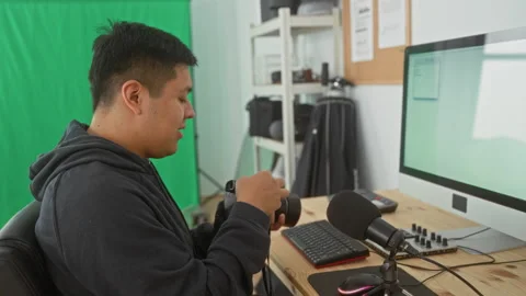 Man adjusting camera at studio desk with green screen, computer and microph.. Video stock 337354907