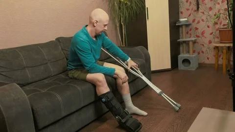 Man adjusting crutches height while sitting on sofa, recovery preparation Stock Footage 333812411