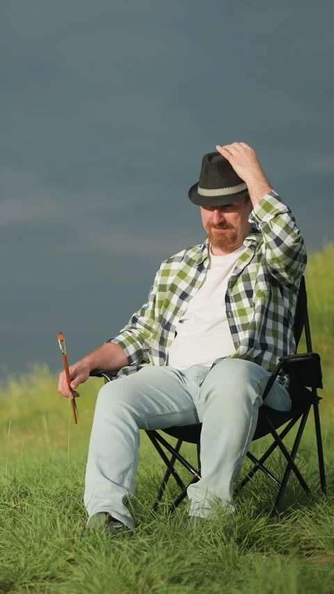 Man Adjusting Hat with Paintbrush in Hand, Sitting in Field under Dark Sky Stock-Footage 306971071