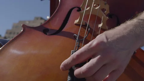 Man adjusting strings on cello outdoors under blue sky by historic buildings Stock Footage 312640139